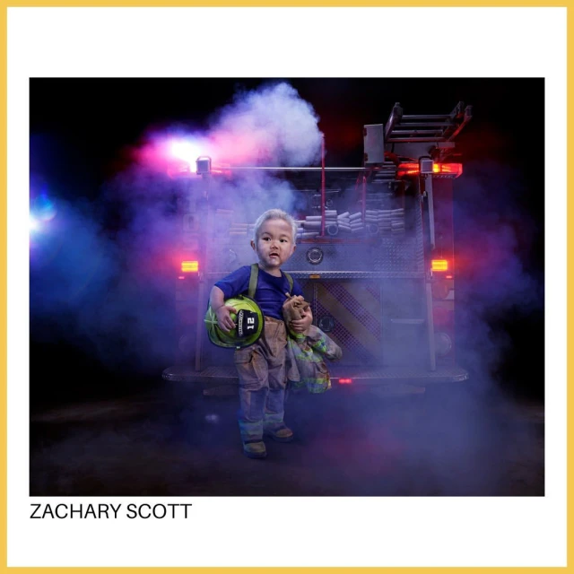 A young child in a firefighter outfit holds a stuffed animal, standing in front of a firetruck in a smoky, dramatic setting.