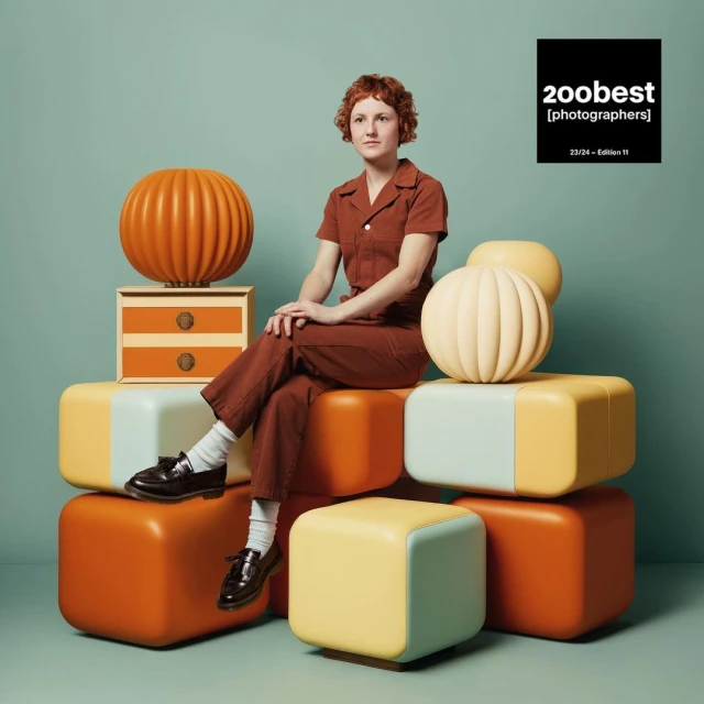 A young woman with short red hair, wearing a brown jumpsuit, sits atop a stack of colorful, rounded cube ottomans in a studio setting.