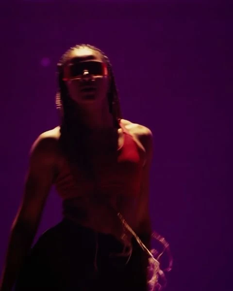 Woman with glasses, top, and skirt stands in purple-toned lighting. She is angled away from the camera and is not looking at the camera.