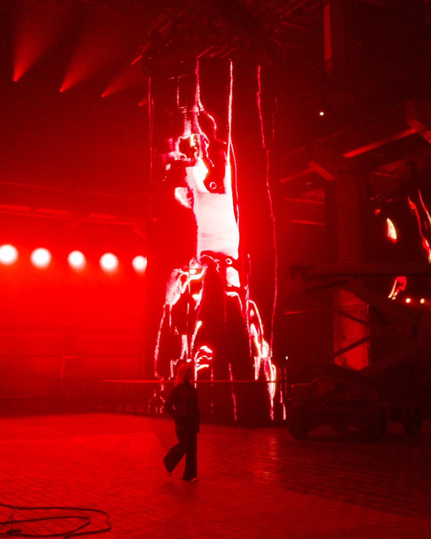 Figure stands on a red floor with a large lit display in the background, illuminated with red spotlights in a red environment.