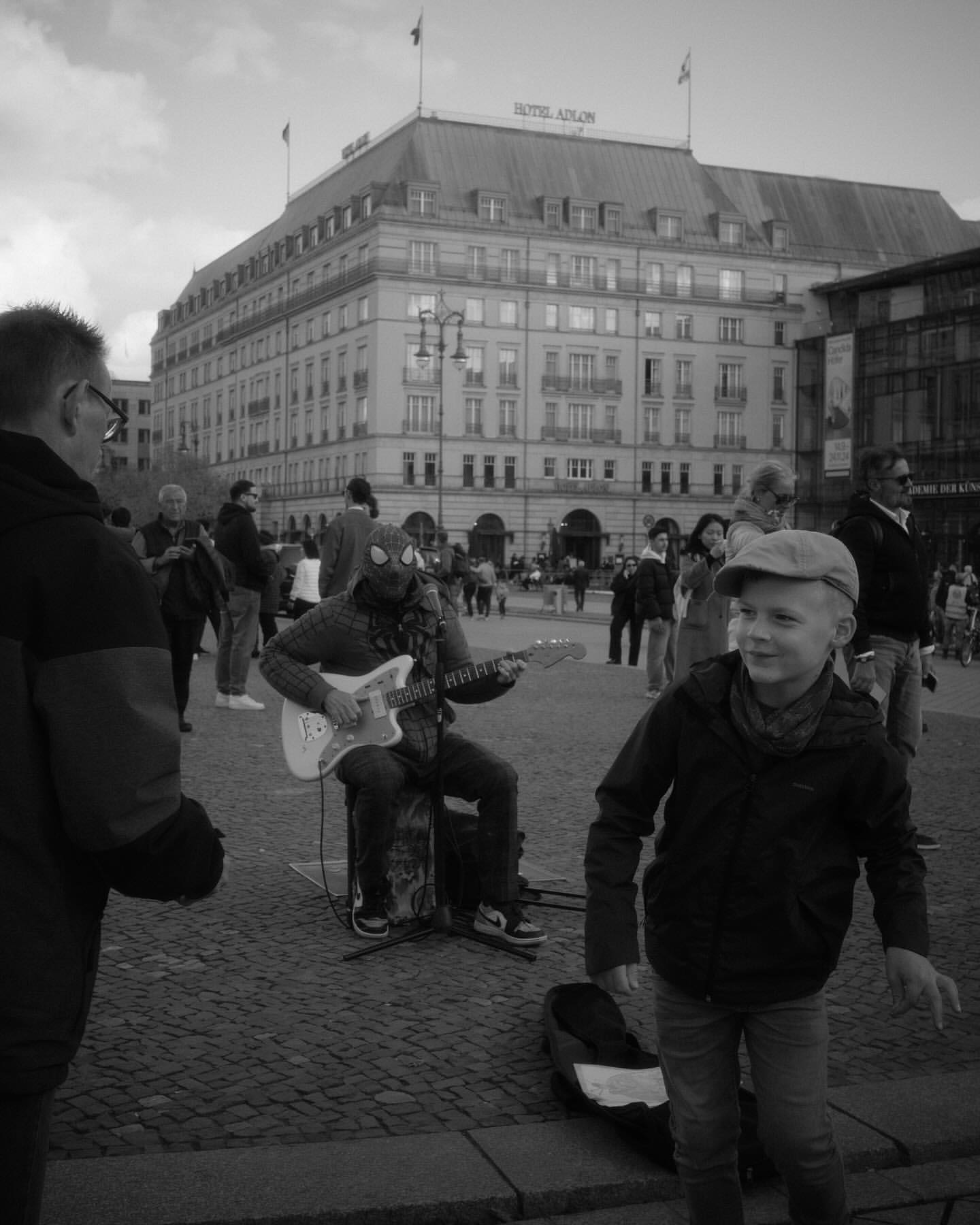 Black and white shot of a musician playing guitar outdoors, with a child looking on and a large building in the background.