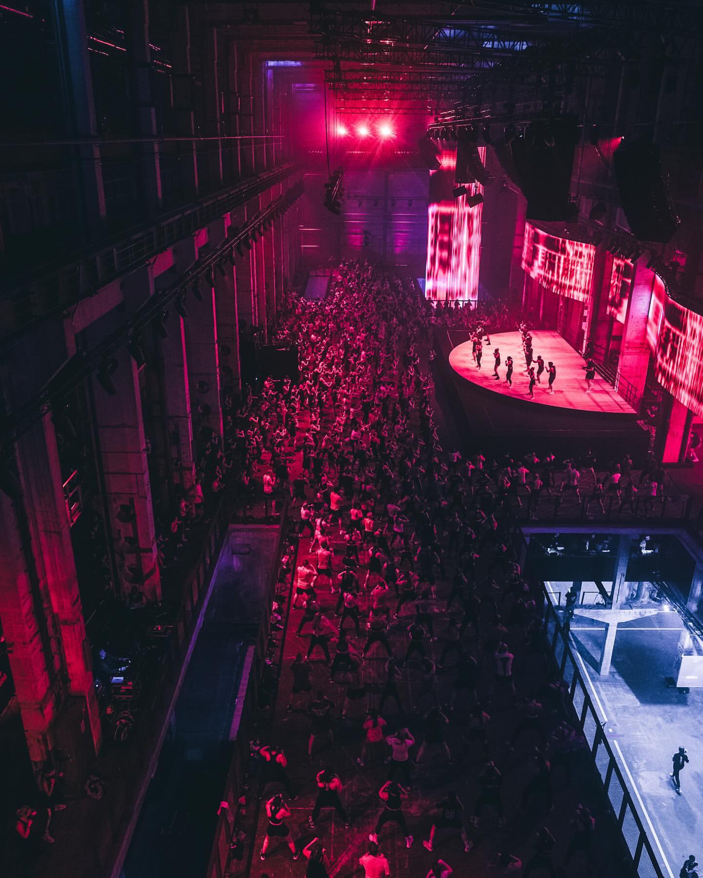 High-angle shot of a large crowd in a spacious industrial venue, illuminated with red and blue lights, with performers on stage.