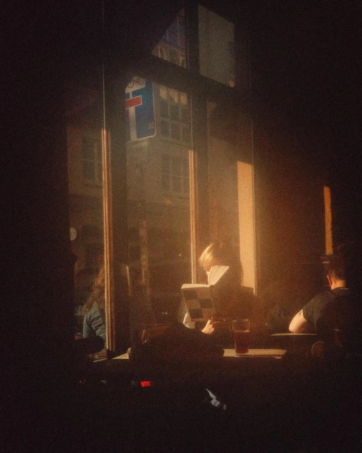 Patrons in a dimly lit cafe setting by a large window read a book and enjoy a drink, with building reflections visible.