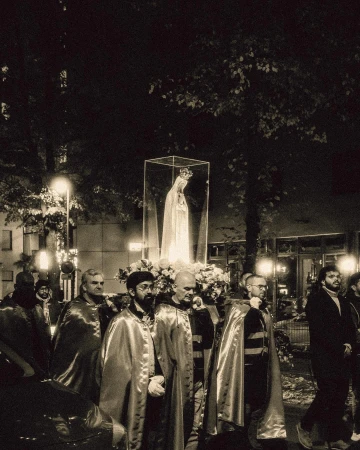 Procession at night with people carrying a statue of the Virgin Mary in a glass case surrounded by white flowers.