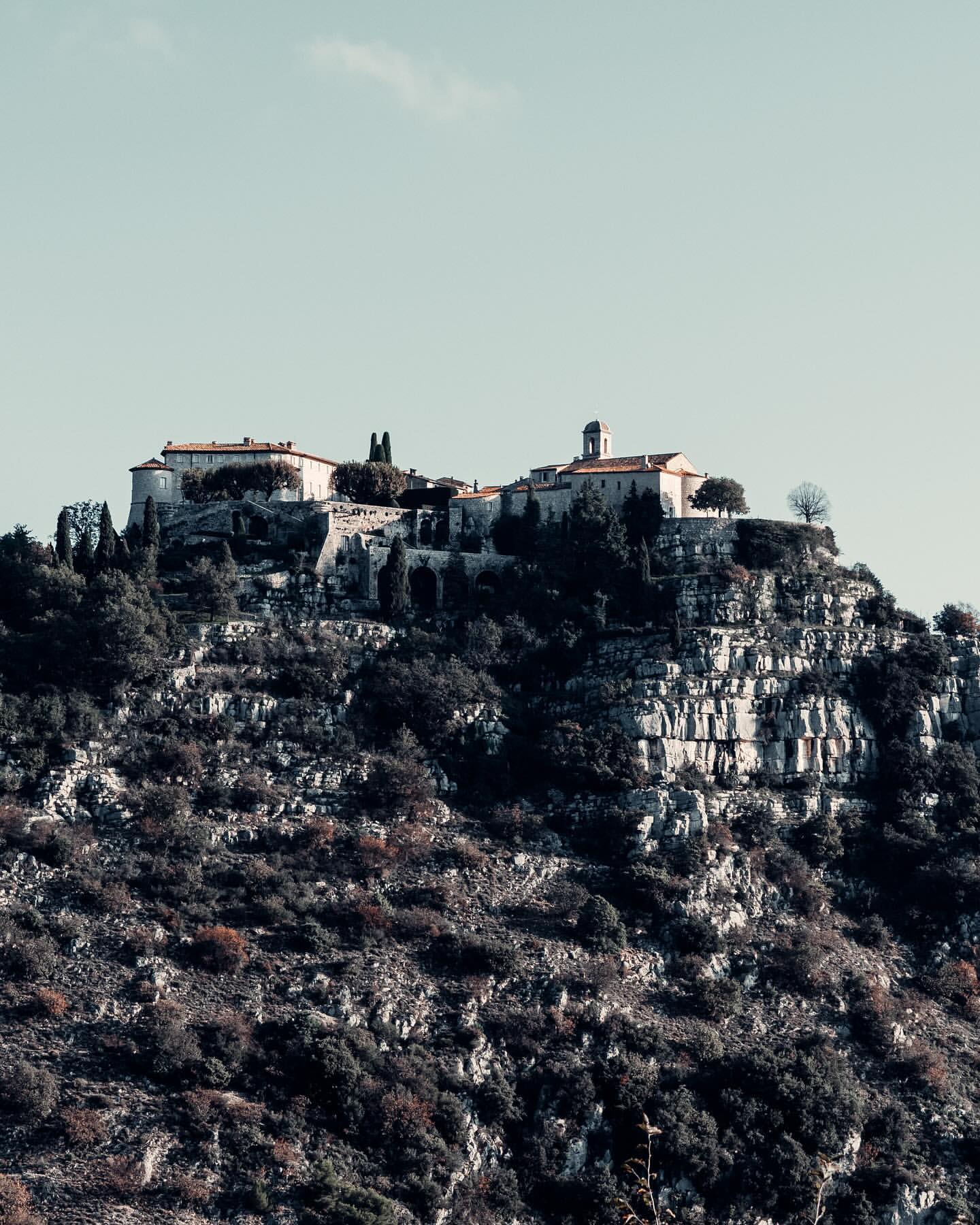 Hilltop village in Eze, France with stone buildings, tile roofs, and lush vegetation on a rocky terrain under a clear sky.