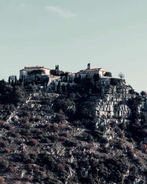 Hilltop village in Eze, France with stone buildings, tile roofs, and lush vegetation on a rocky terrain under a clear sky.