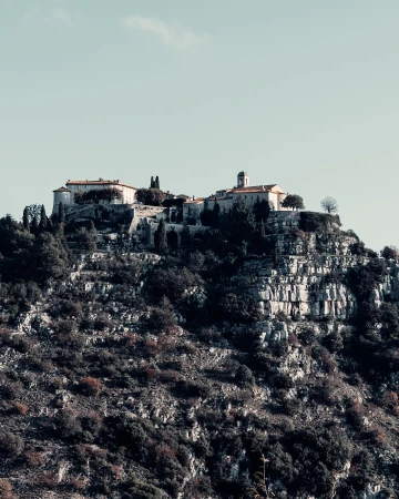 Hilltop village in Eze, France with stone buildings, tile roofs, and lush vegetation on a rocky terrain under a clear sky.