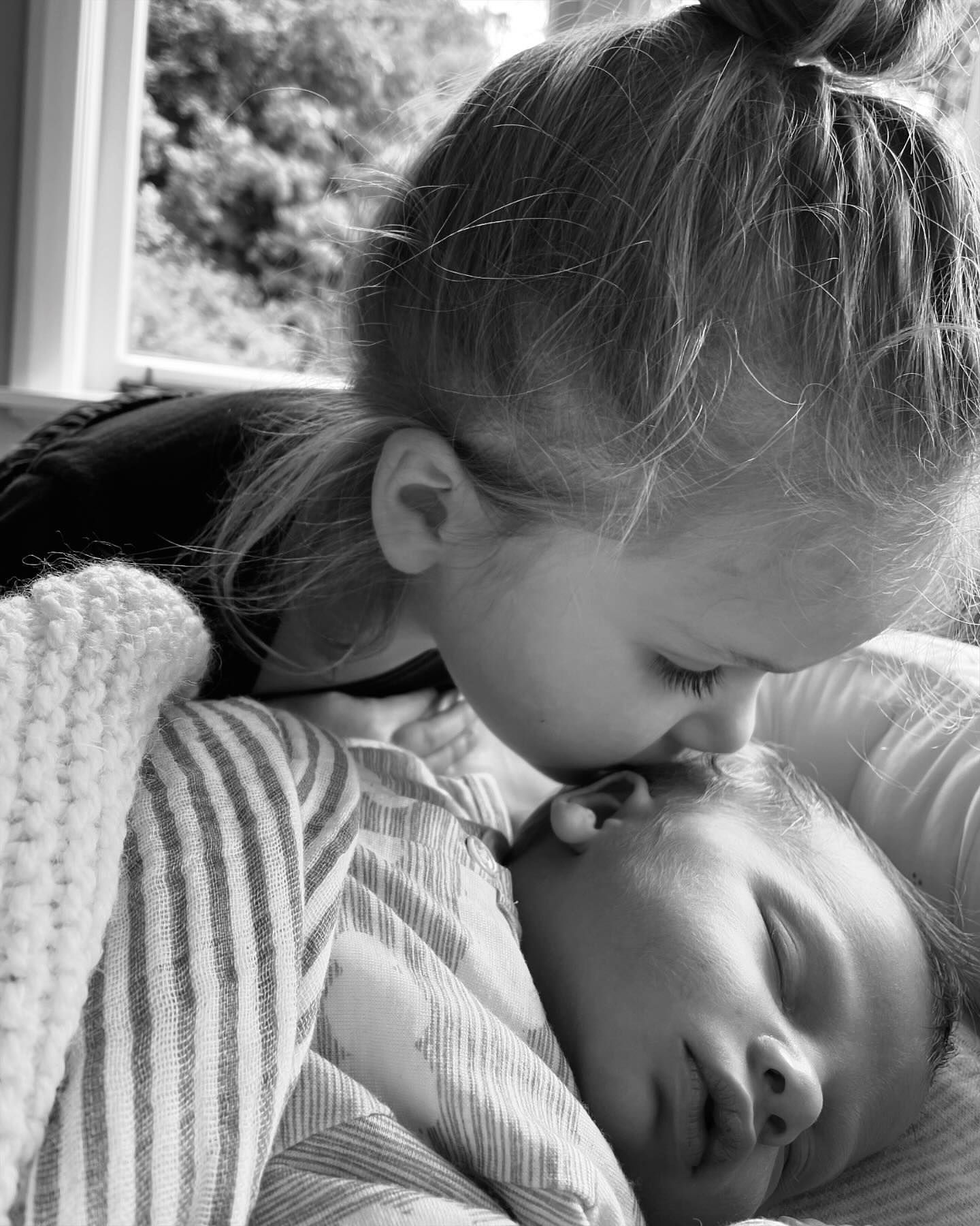 Black and white portrait of a young girl kissing a sleeping baby's forehead, capturing a tender sibling moment indoors.