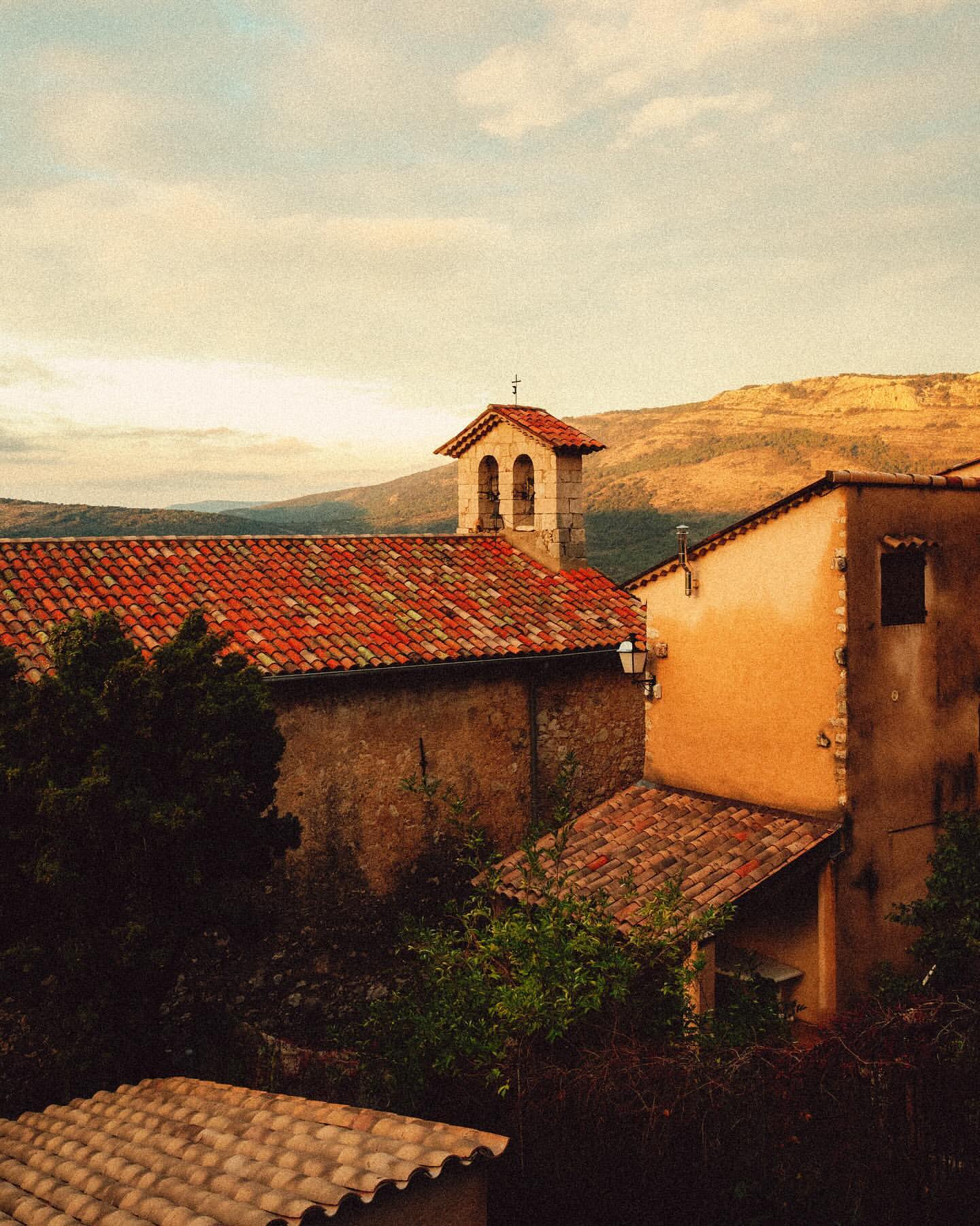 Tiled roofs of old buildings and church tower in a rural European town, with mountains in the background against a cloudy sky.