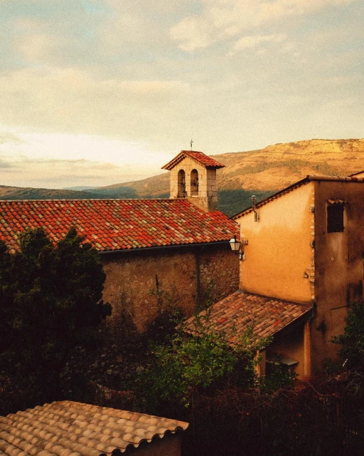 Tiled roofs of old buildings and church tower in a rural European town, with mountains in the background against a cloudy sky.