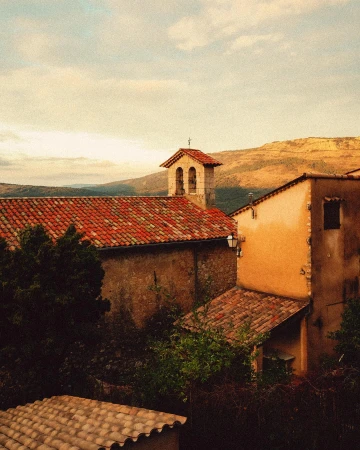 Tiled roofs of old buildings and church tower in a rural European town, with mountains in the background against a cloudy sky.