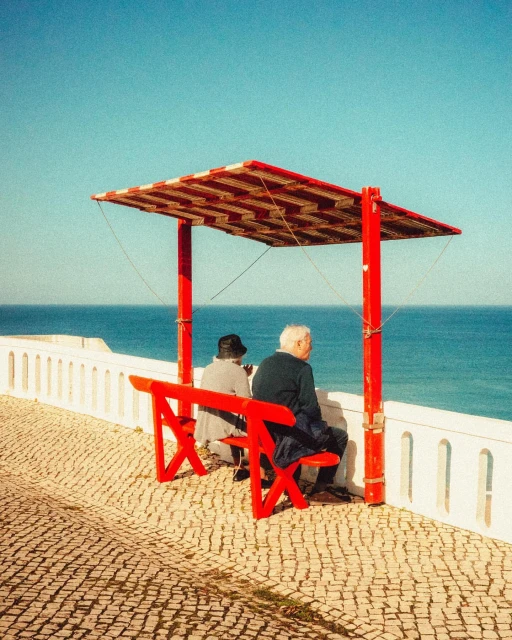 Two elderly adults sit on a red bench under a shelter overlooking the ocean on a sunny day. The scene evokes a sense of peace and solitude.