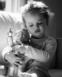 Monochrome portrait of a child hugging a small figurine, seated on a sofa with natural side lighting.