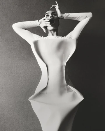 Model in architectural white sculptural dress with hands raised to face, photographed in monochrome.