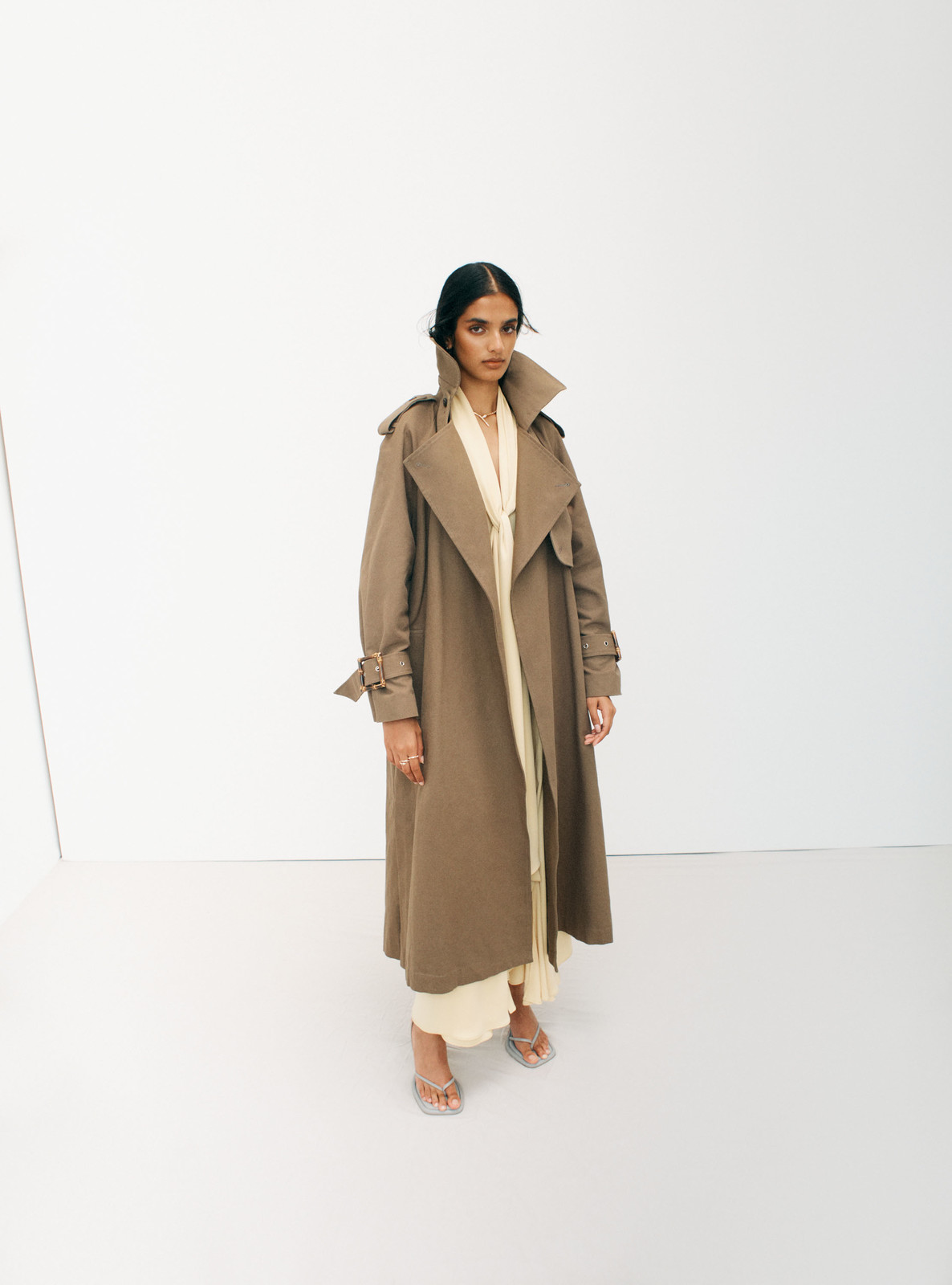 Model wearing a long brown trench coat over cream attire against a plain white studio background.