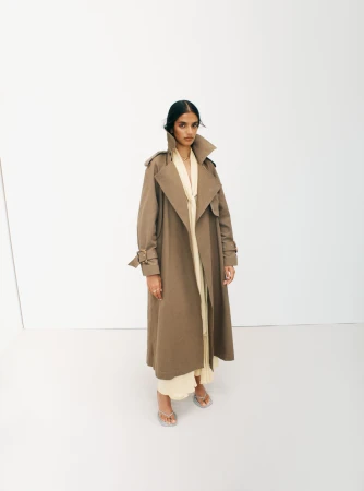 Model wearing a long brown trench coat over cream attire against a plain white studio background.