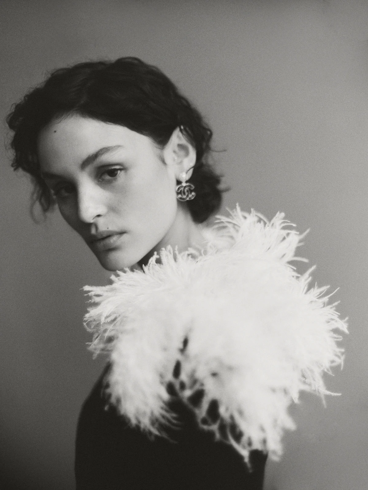 Portrait of a stylish woman in a black top with white feathered collar and Chanel earrings, set against a neutral gray background.
