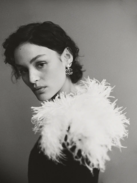 Portrait of a stylish woman in a black top with white feathered collar and Chanel earrings, set against a neutral gray background.