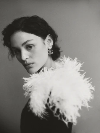 Portrait of a stylish woman in a black top with white feathered collar and Chanel earrings, set against a neutral gray background.