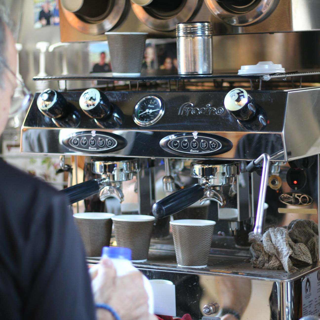 An indoor shot shows a barista preparing coffee on a shiny espresso machine, with paper cups and a milk bottle visible.