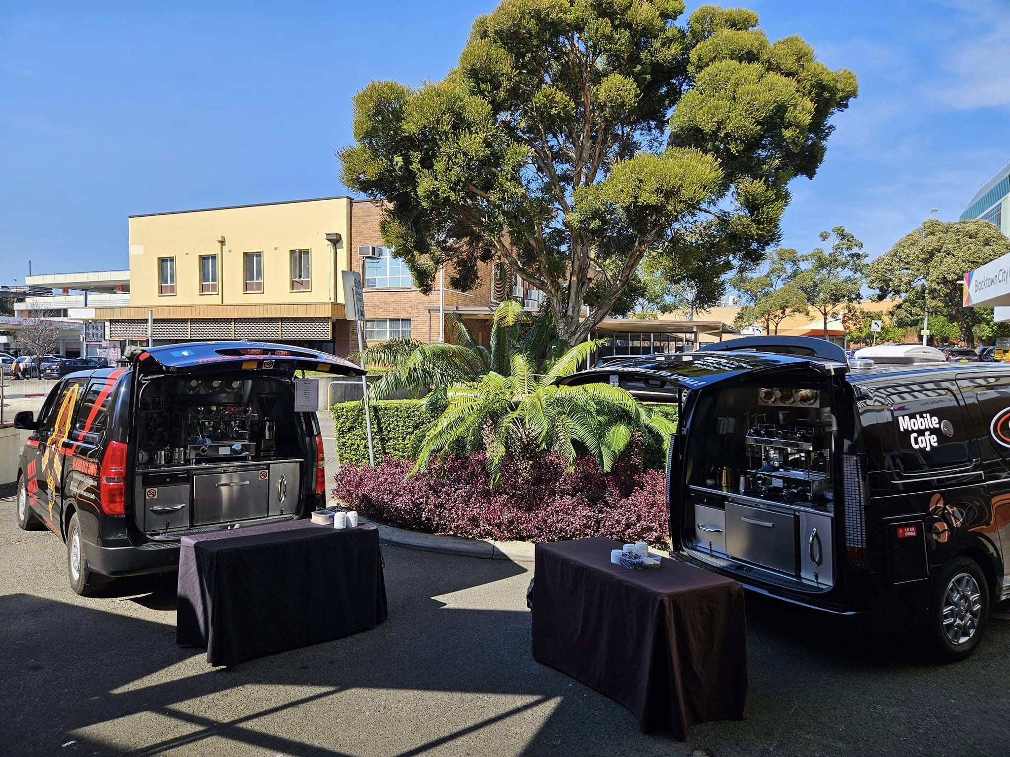 Two black mobile cafe vans parked outdoors with tables and cups on display, set against a backdrop of trees and buildings under a blue sky.
