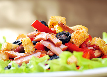 Salad featuring lettuce, sliced ham, peppers, olives, croutons, and dressing on a white plate, with a neutral background.
