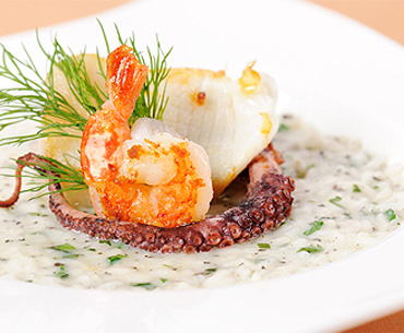 A prepared dish featuring a cooked shrimp, a piece of fish, and an octopus tentacle on a bed of risotto, presented on a white plate.