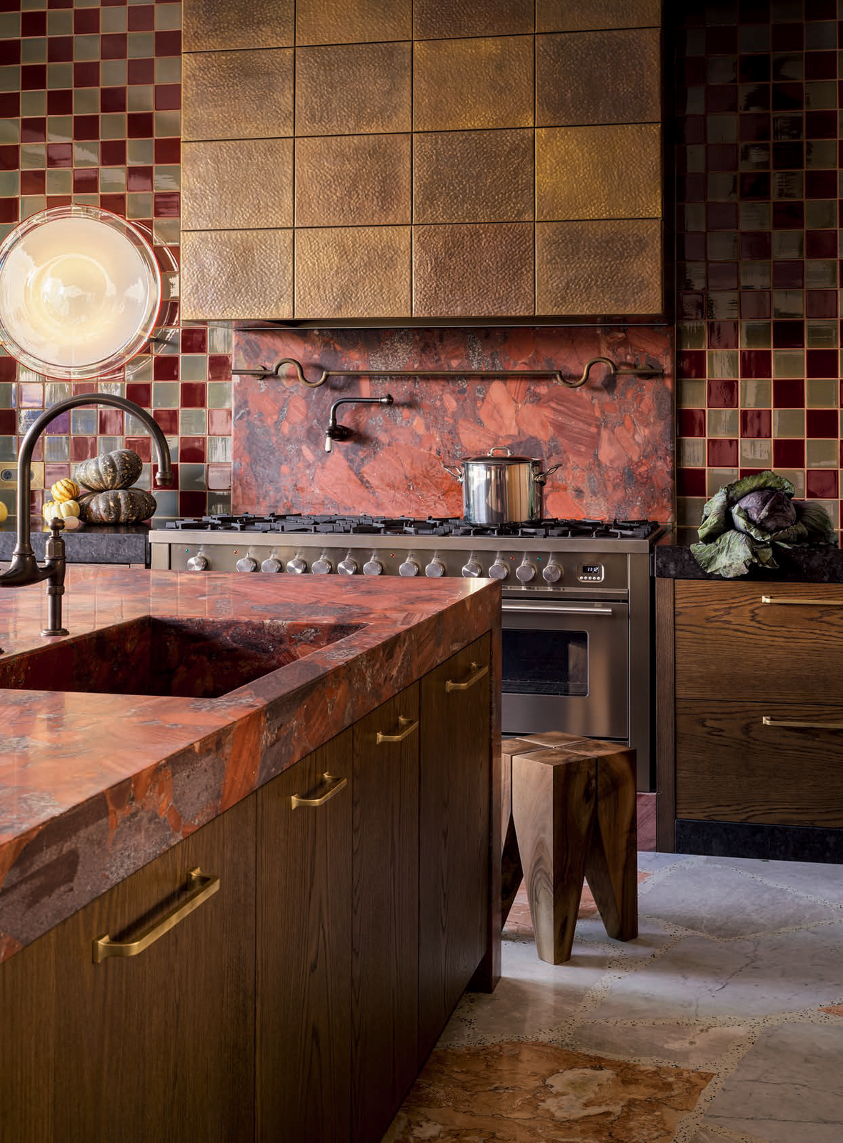 Warm-toned kitchen interior featuring a kitchen island with a sink, an oven range with a pot, a wooden stool, and a checkered backsplash.
