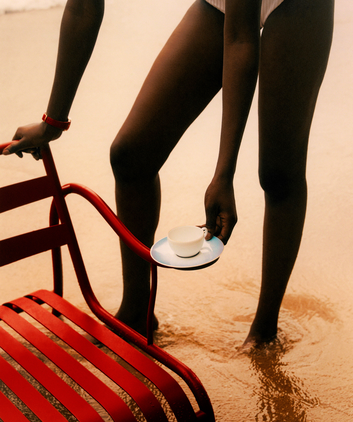 Woman in white swimsuit standing on beach next to red chair, holding saucer with cup, minimalist and evocative summer scene, relaxation.