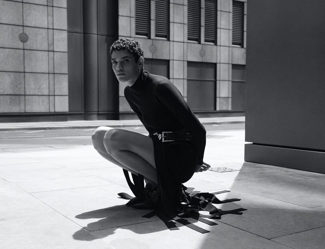 Monochrome portrait of a person crouching on stone pavement wearing a black turtleneck and dark skirt.