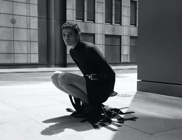Monochrome portrait of a person crouching on stone pavement wearing a black turtleneck and dark skirt.