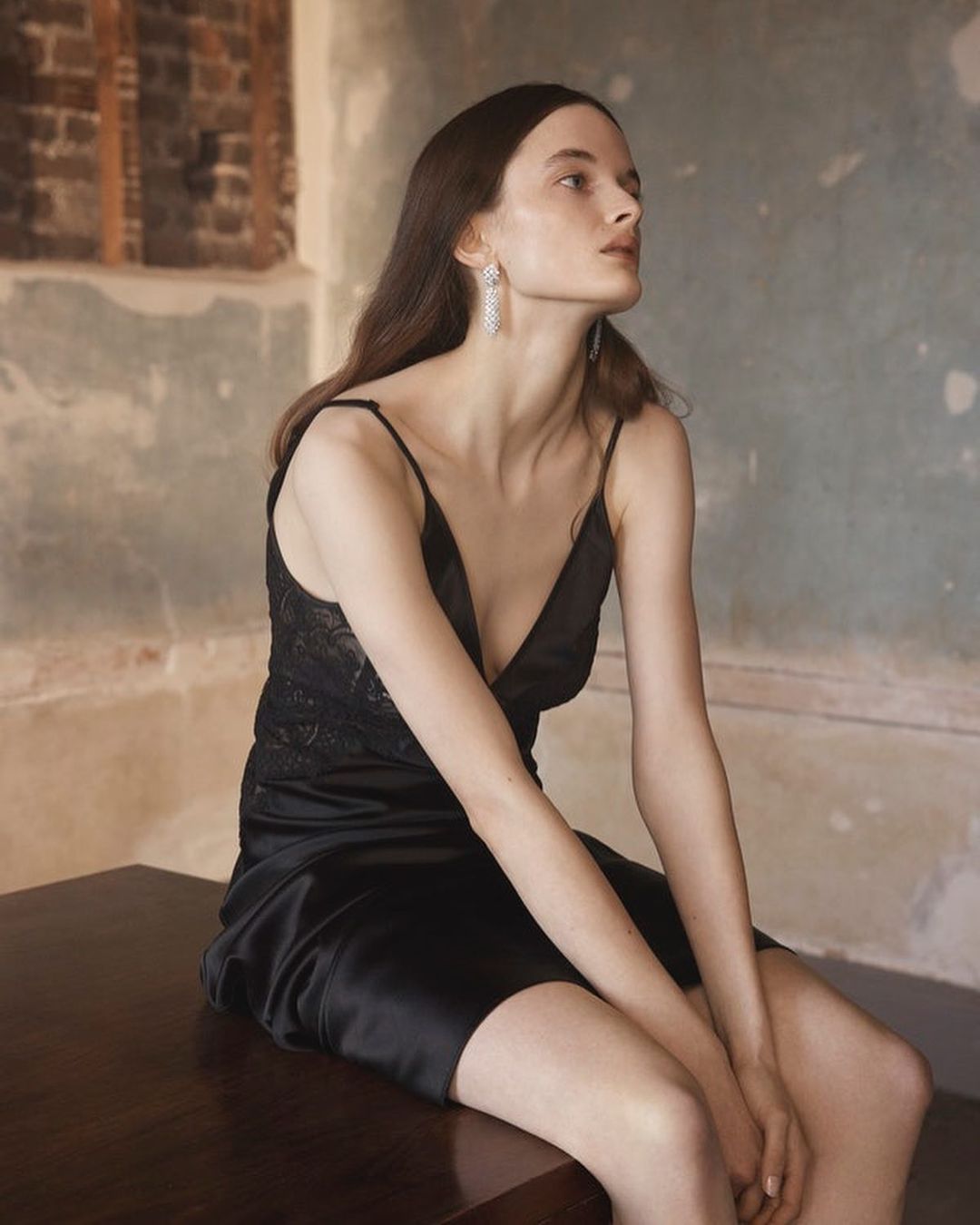 Elegant woman in a black lace and silk slip dress poses indoors with soft lighting, showcasing a blend of modern fashion and classic style.