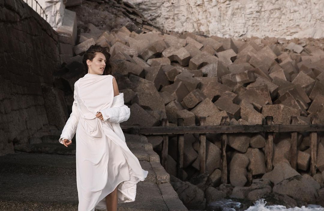 Adult female in a long white dress poses on a stone path near a rocky coastline with piled stone cubes and ocean water.