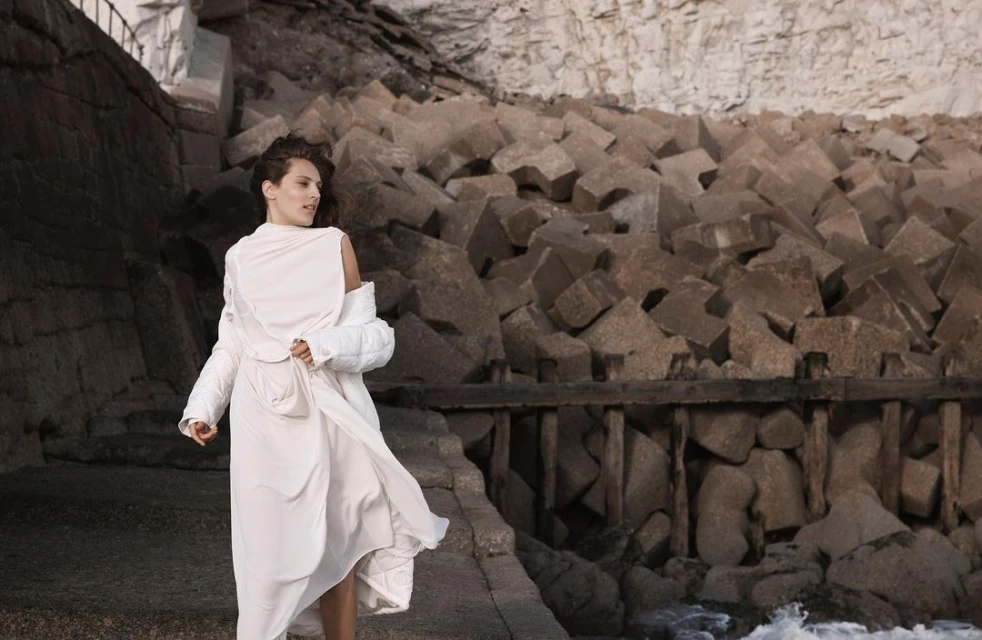 Adult female in a long white dress poses on a stone path near a rocky coastline with piled stone cubes and ocean water.