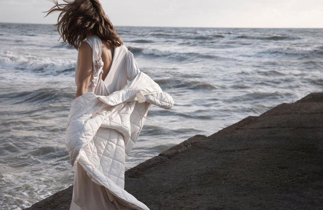 Back view of woman in dress with jacket standing on concrete steps by the ocean with waves and breeze on a cloudy day.