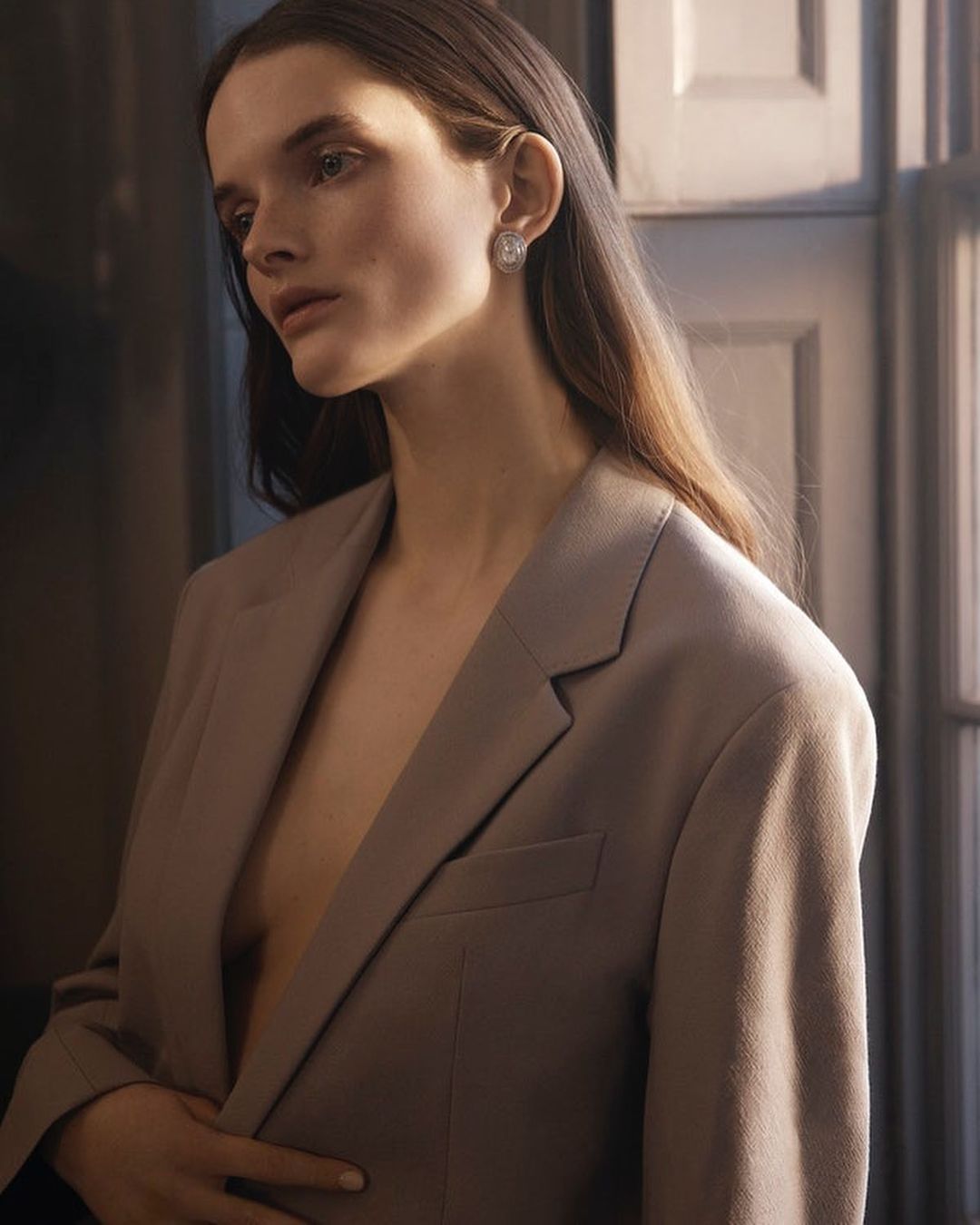 Portrait of a woman with long brown hair wearing a beige blazer and silver earrings, captured in a dimly lit indoor setting.