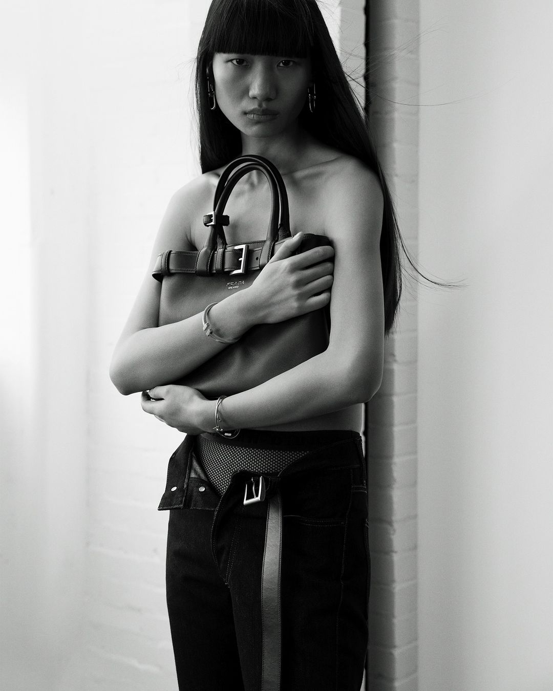 Asian woman in black denim with open waist holding a brown leather handbag in a black-and-white fashion studio portrait.