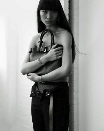 Asian woman in black denim with open waist holding a brown leather handbag in a black-and-white fashion studio portrait.