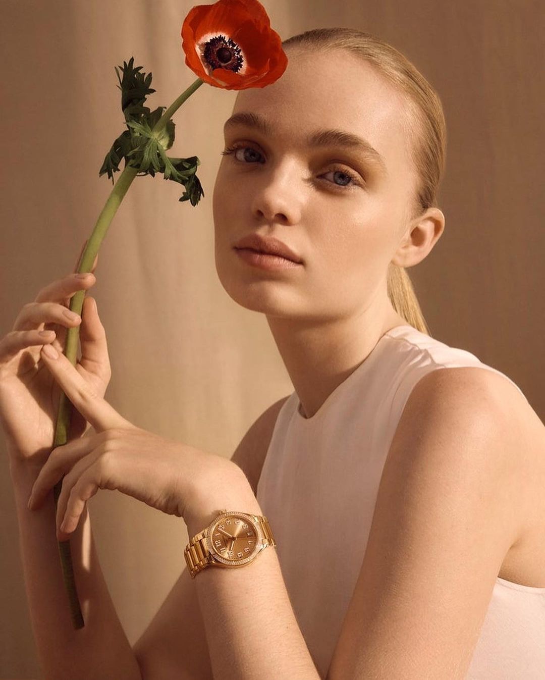 Blond woman in a white top poses with a red flower and wears a gold watch against a tan backdrop in a medium studio shot.