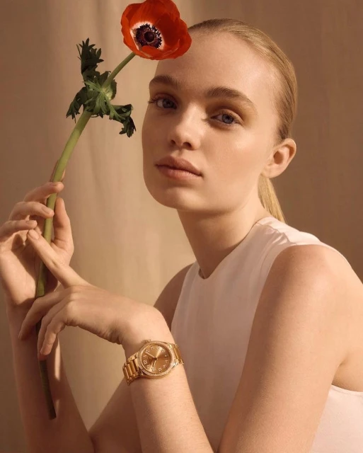 Blond woman in a white top poses with a red flower and wears a gold watch against a tan backdrop in a medium studio shot.