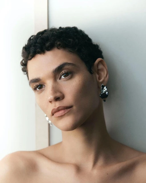 Close-up portrait of a young adult woman with short curly dark hair and drop earrings looking at the camera indoors, against a neutral wall.