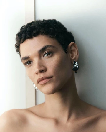 Close-up portrait of a young adult woman with short curly dark hair and drop earrings looking at the camera indoors, against a neutral wall.