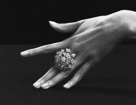 A woman's hand elegantly displays an ornate ring with gemstone detailing against a stark black background in this black and white studio shot.