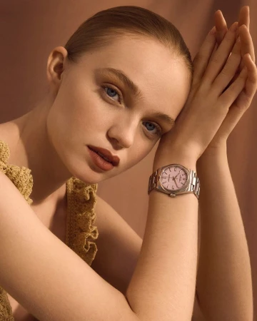 Elegant studio shot of a young woman wearing a watch with a silver band, with her hands near her face and a soft, neutral backdrop.