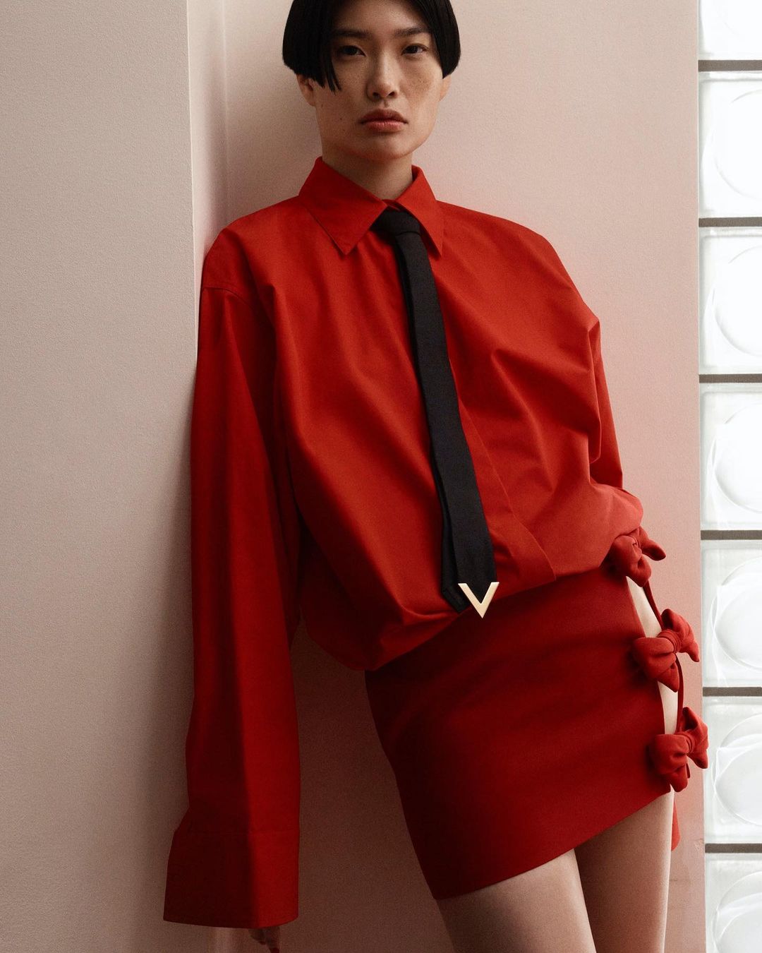 Stylish woman in a red oversized shirt, black tie, and red mini skirt with bows poses against a neutral wall and window.