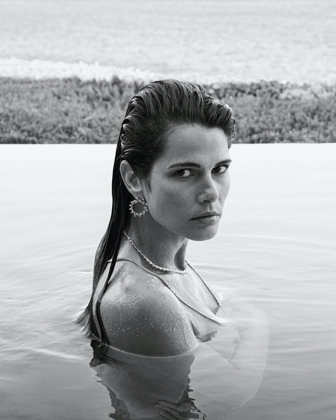 Monochrome portrait of an adult woman in water, wearing jewelry; a tranquil outdoor scene with a natural background and a soft, desaturated palette.