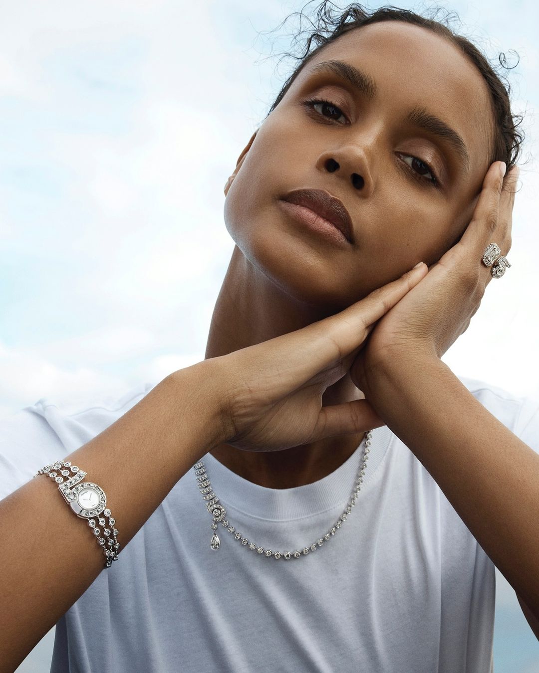 A woman with tan skin poses outdoors, wearing a white T-shirt and sparkling jewelry: watch, ring, and necklace, against a cloudy sky.