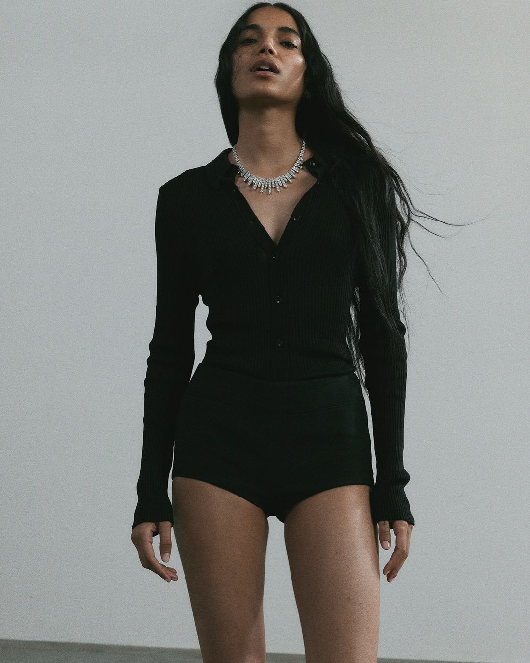 Elegant shot of a woman with long hair wearing a black collared shirt and shorts, accessorized with a diamond necklace against gray backdrop.