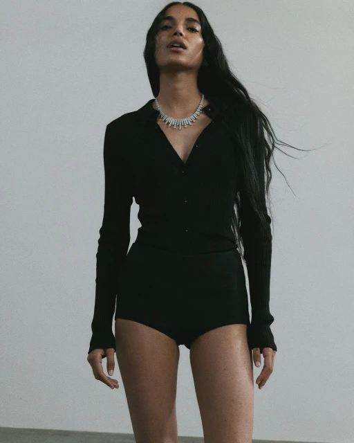 Elegant shot of a woman with long hair wearing a black collared shirt and shorts, accessorized with a diamond necklace against gray backdrop.