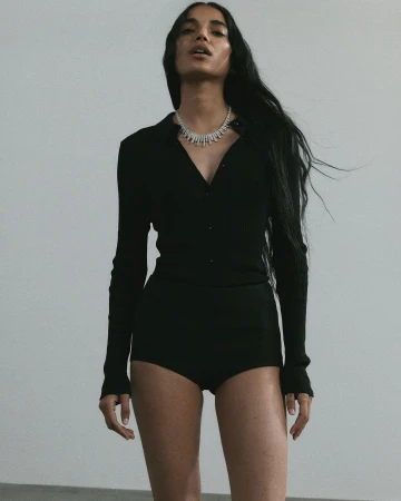 Elegant shot of a woman with long hair wearing a black collared shirt and shorts, accessorized with a diamond necklace against gray backdrop.